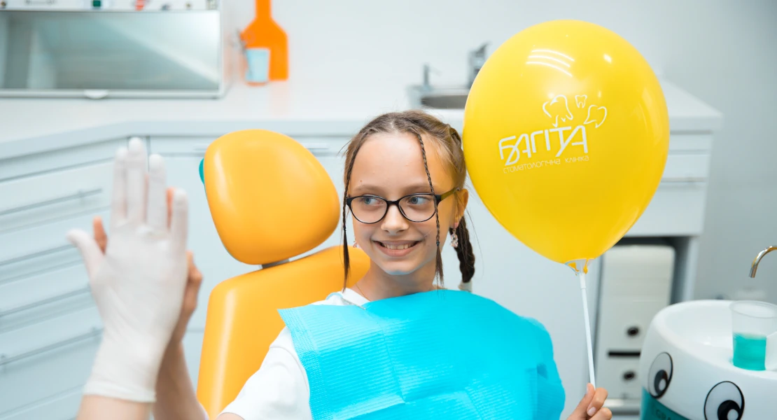 Лікування зубів дівчинці в дитячому кабінеті, у стоматологічній клініці Багіта в Черкасах, вона усміхаючись дає лікарю п'ять і тримає в руках фірмову жовту кульку з написом Багіта