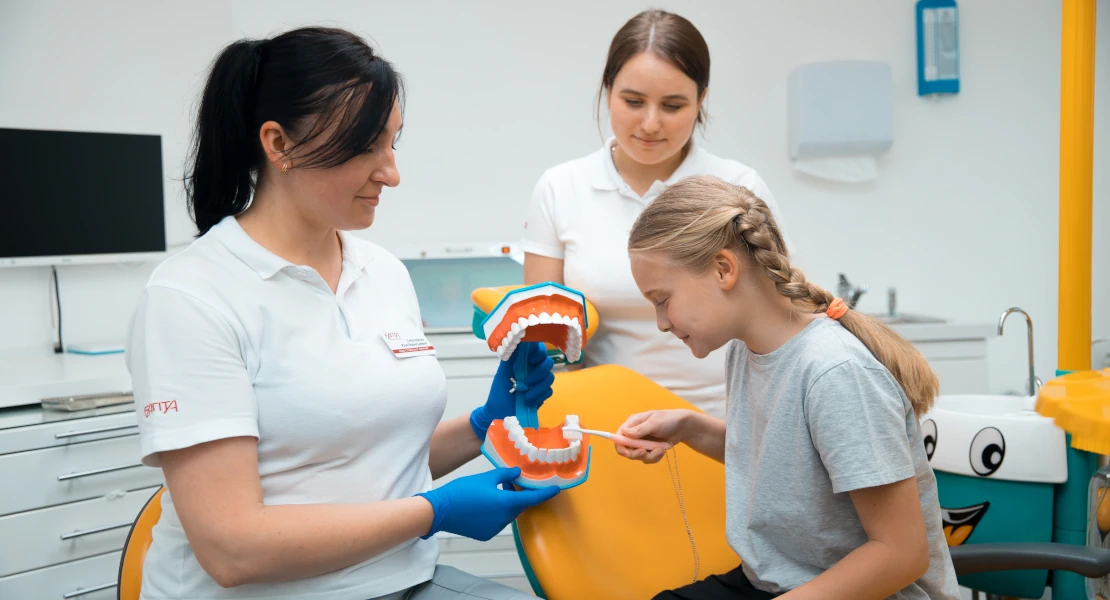 на фото ребенок в детском стоматологическом кабинете рассматривает с врачем муляж челюсти и зубной щёткой учится правильно чистить зубы